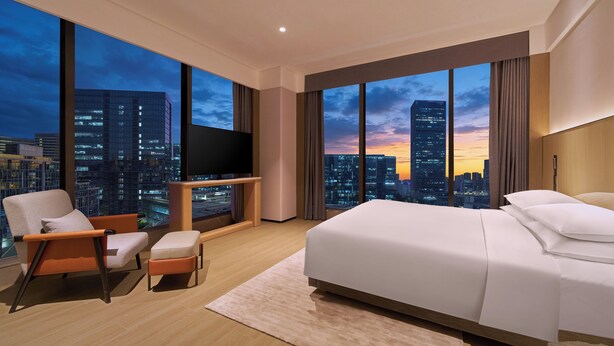 A modern hotel room with a large bed, a TV, a chair, and a view of a cityscape at sunset. - Hyatt Place Shenzhen Longgang (Shenzhen)