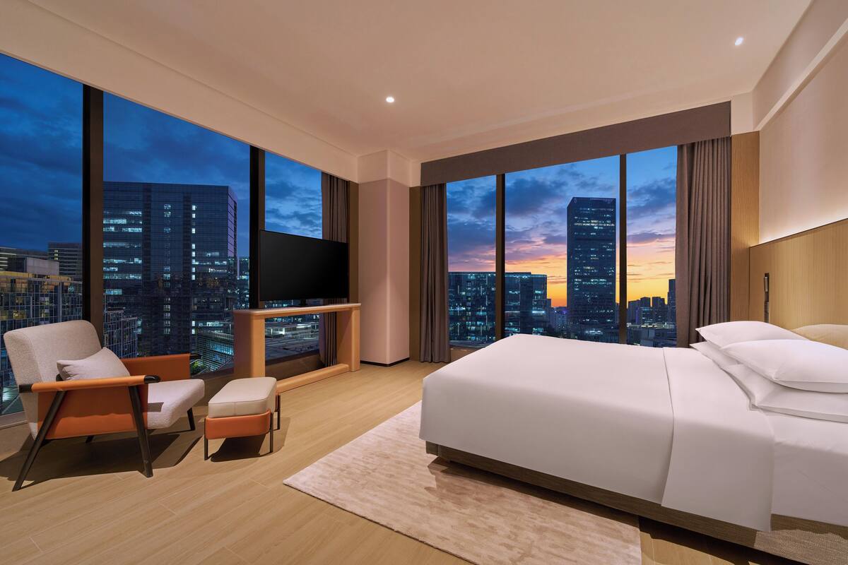 Een moderne hotelkamer met een groot bed, een televisie, een stoel en uitzicht op een stadslandschap bij zonsondergang.