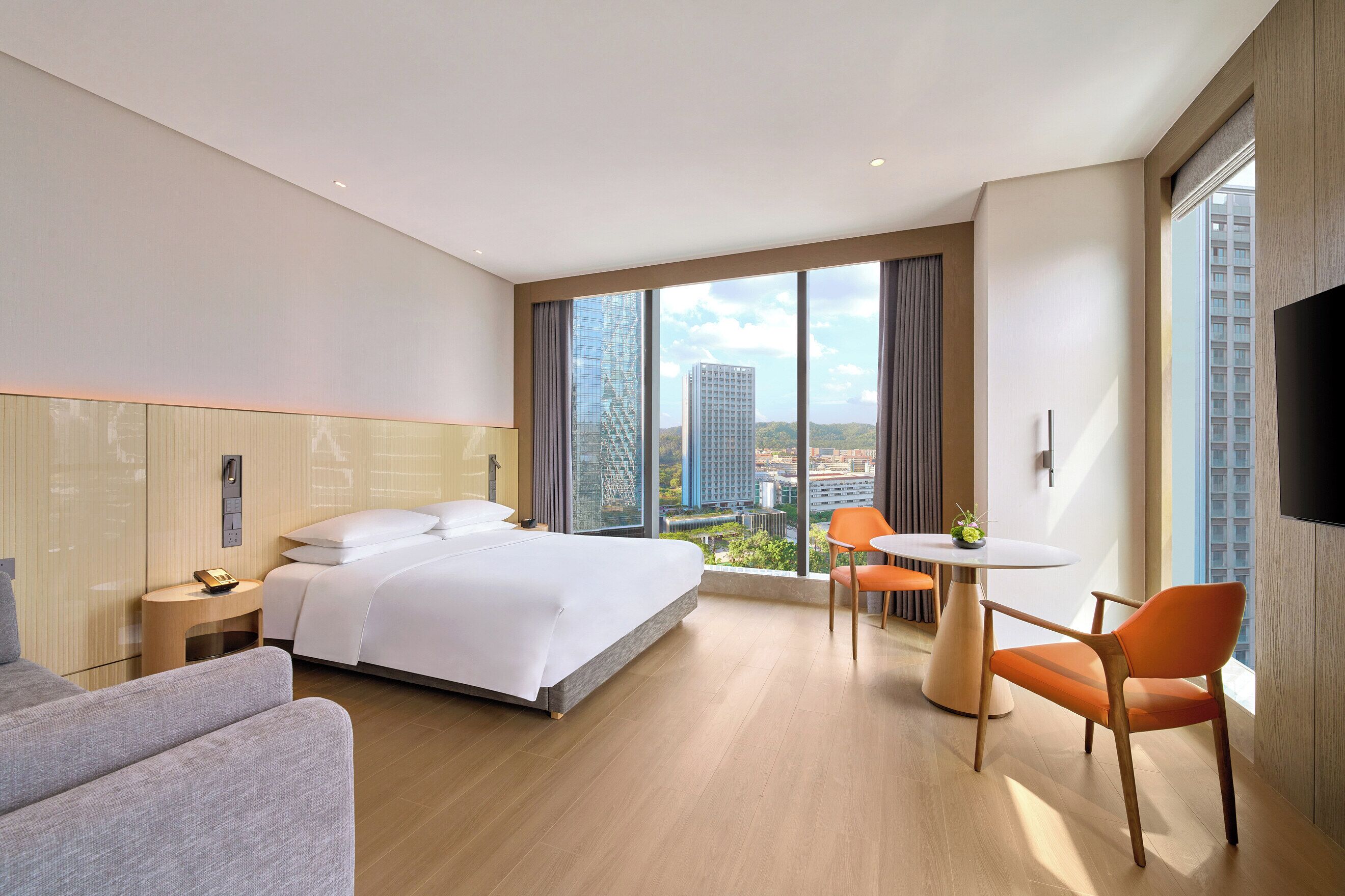Habitación de hotel moderna con cama, escritorio y silla, un amplio ventanal con vista a la ciudad y un televisor de pantalla plana.