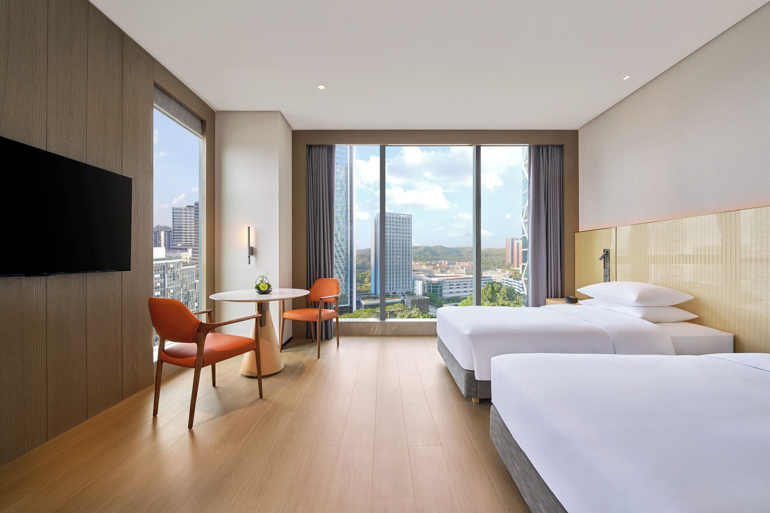 Habitación de hotel con dos camas, televisor, zona de comedor con sillas y un amplio ventanal con vista a la ciudad.