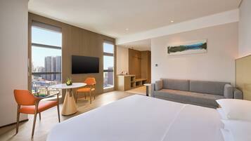Habitación de hotel moderna con una cama grande, un sofá, una mesa de comedor con sillas, un televisor y vistas a la ciudad.