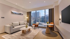 A modern hotel room with a sofa, two coffee tables, a dining table with chairs, a large window with a city view, and a flat-screen TV mounted on the wall. - Hyatt Place Shenzhen Longgang (Shenzhen)