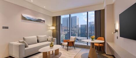 A modern hotel room with a sofa, two coffee tables, a dining table with chairs, a large window with a city view, and a flat-screen TV mounted on the wall.