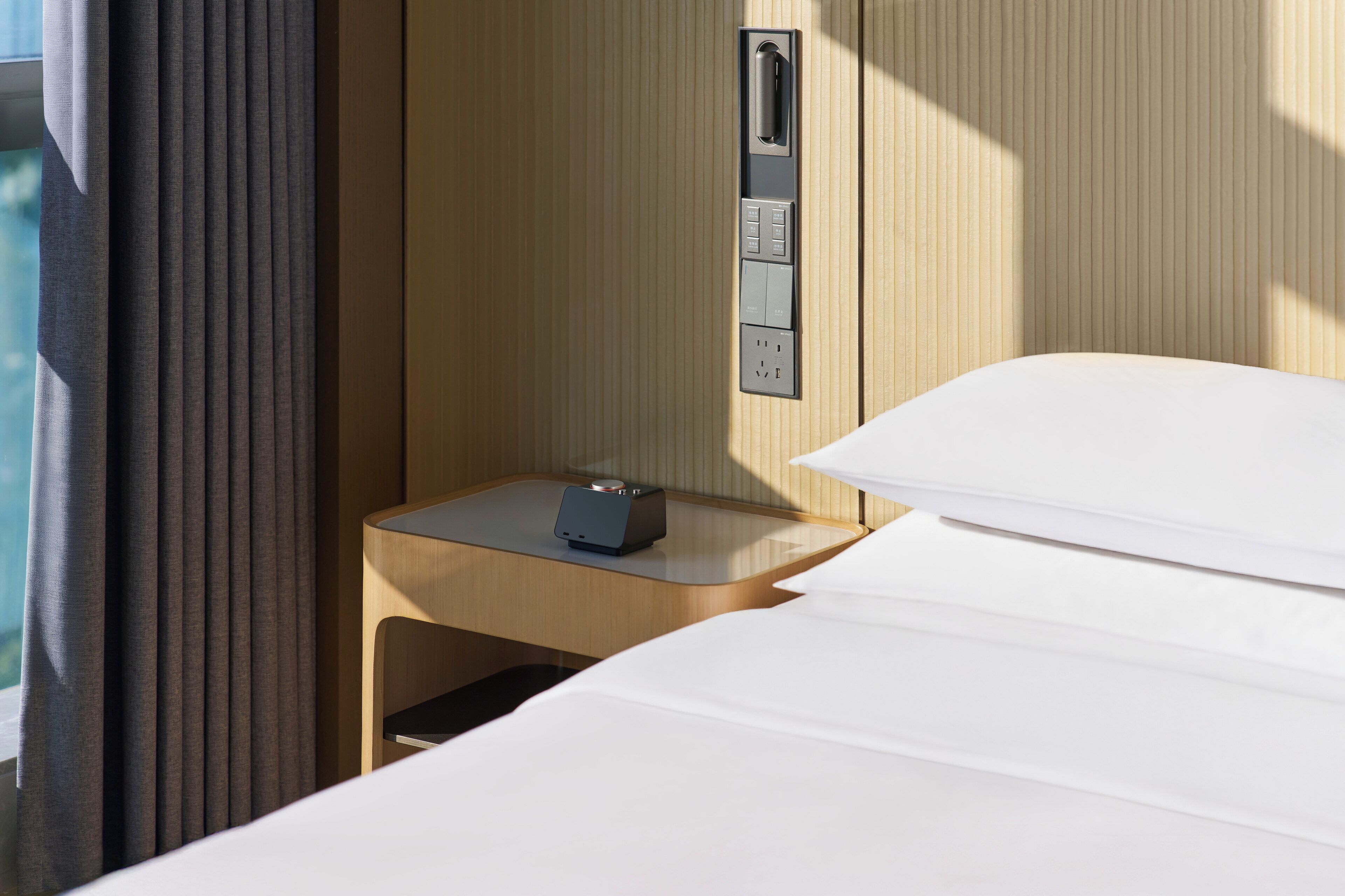 A hotel room with a bed, a bedside table with a clock, a power outlet, and a wall-mounted control panel.