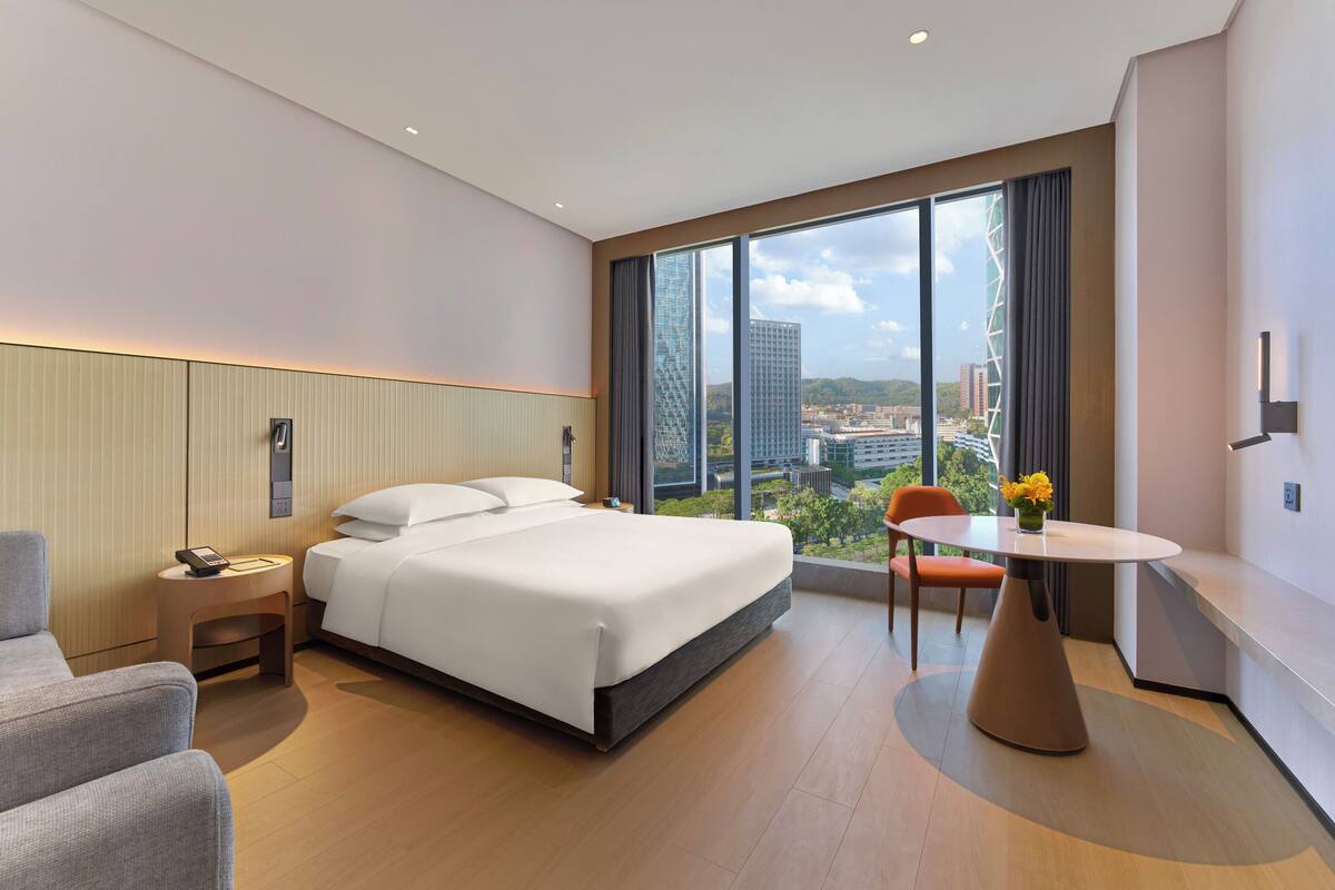 Een moderne hotelkamer met een groot bed, een klein tafeltje, een stoel en een uitzicht op de skyline door grote ramen.