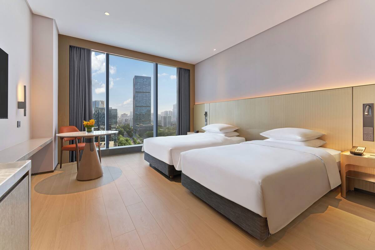 Een moderne hotelkamer met twee bedden, een bureau, een stoel en een groot raam met uitzicht op de stad.