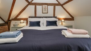 A bedroom with a high ceiling, exposed wooden beams, a large bed with two pillows, bedside tables with lamps, and a framed picture on the wall. - Magpie Cottage (Worcester)