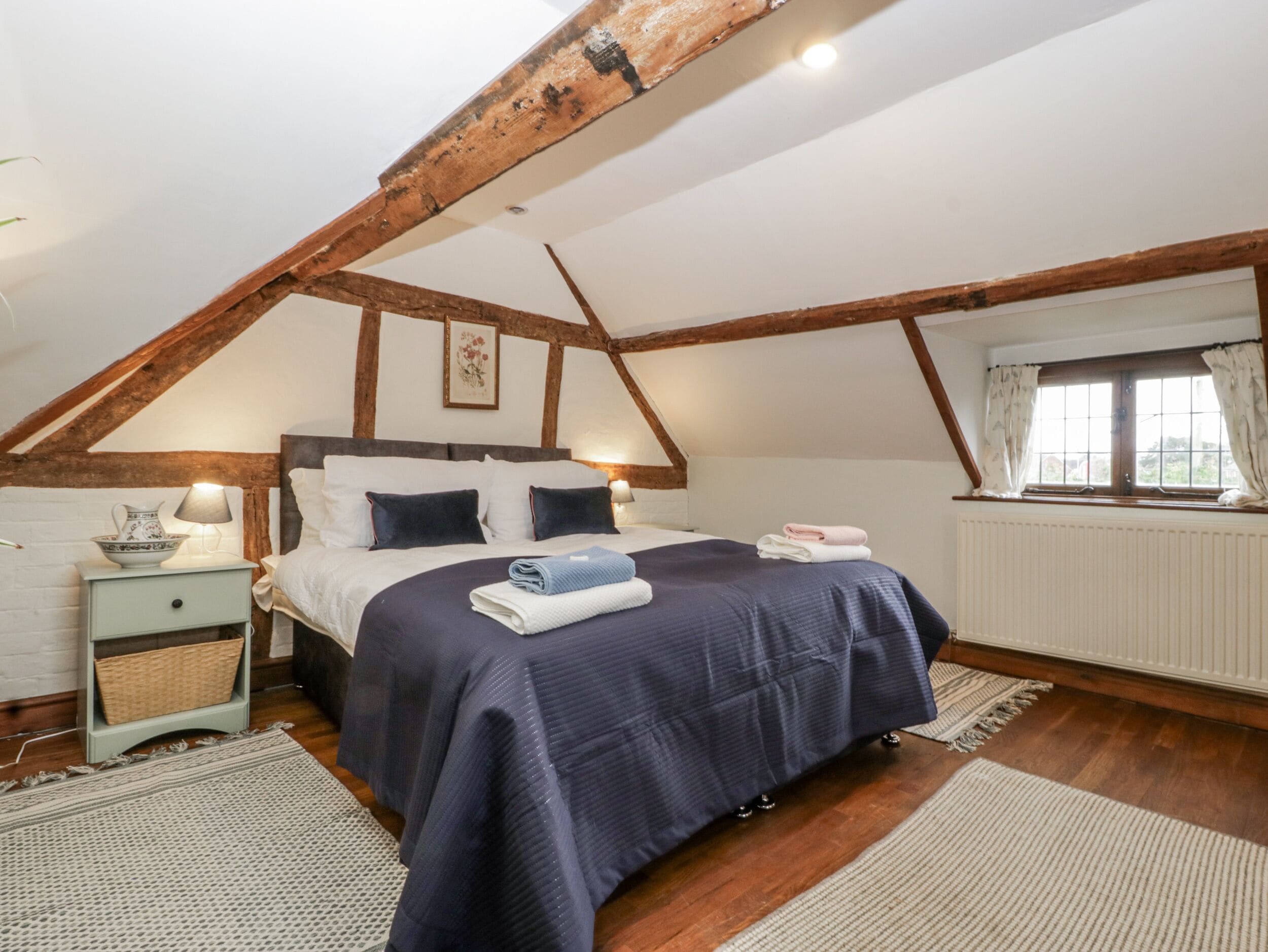 A bedroom with a sloped ceiling, a bed with a dark blue bedspread, a nightstand with a lamp, and a window with curtains.