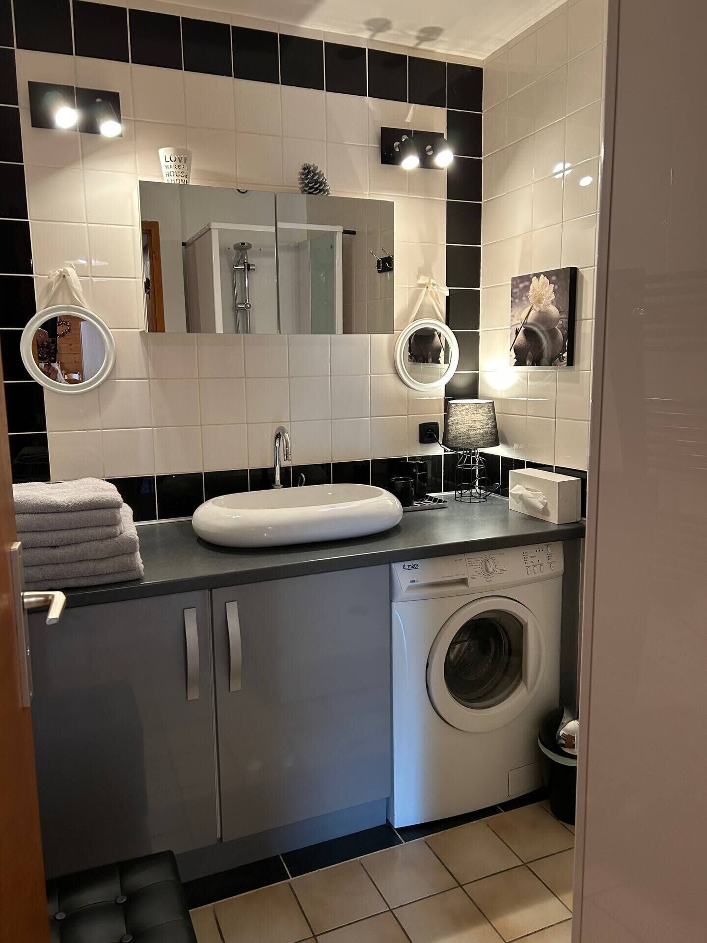 A compact bathroom with a white sink, two wall-mounted mirrors, a black countertop, a washing machine, and a towel rack.