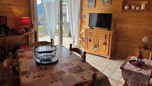 Acogedor interior de cabaña de madera con mesa de comedor, sillas, televisor y zona de cocina con estantes y un armario.