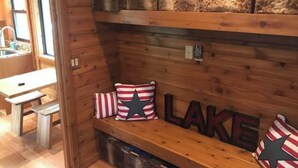Interior de una cabaña de madera con un banco, almohadones y un estante en la pared con frascos.