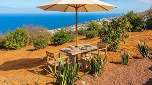 Un espacio de comedor al aire libre con mesa, sillas y una sombrilla grande, con vistas a un paisaje costero con casas y vegetación.