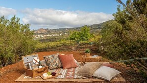 An outdoor seating area with a view of a hilly landscape, a picnic setup with food and drinks, and a small table with a book. - Ursula's Cozy Retreat in Santa Ursula (Santa Ursula)