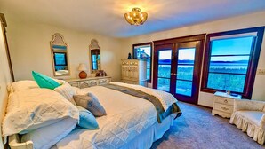 Un dormitorio con una cama grande, un aparador, una mesita de noche y vistas a un bosque por las ventanas.