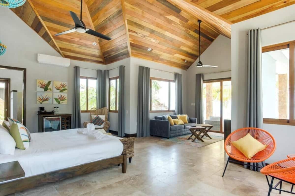 A spacious bedroom with a large bed, a sofa, and a chair. There are windows and a ceiling fan.