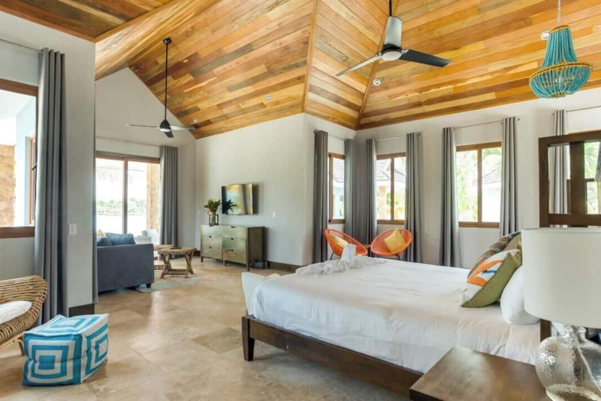 A spacious bedroom with a large bed, a desk, and a chair. There are large windows and a ceiling fan.