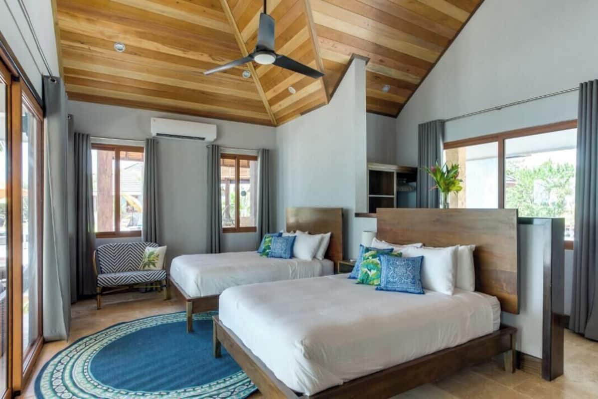 A modern hotel room with two beds, a ceiling fan, large windows, and a view of the outside.