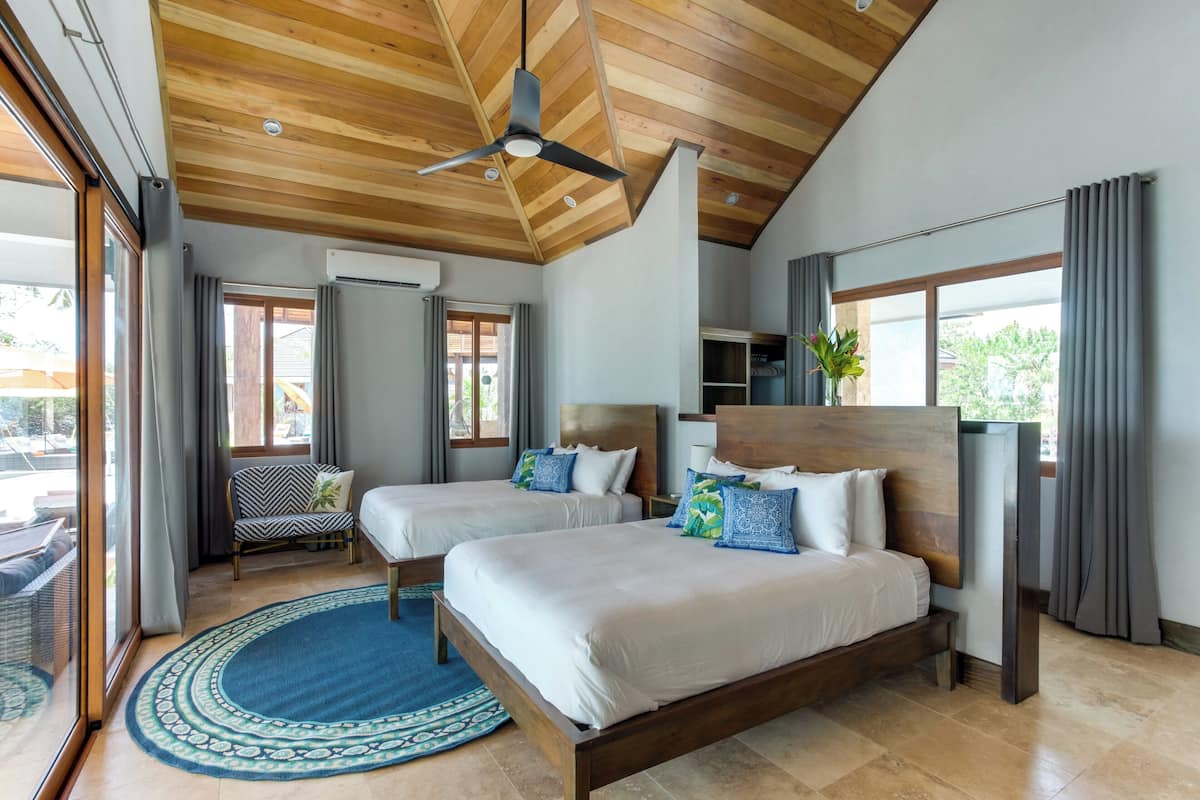 A modern hotel room with two beds, a ceiling fan, large windows, and a view of the outside.
