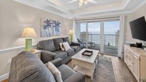 A living room with a grey sofa, a coffee table, a TV, and a balcony with a view of the ocean. - Crystal Tower 906 (Gulf Shores)