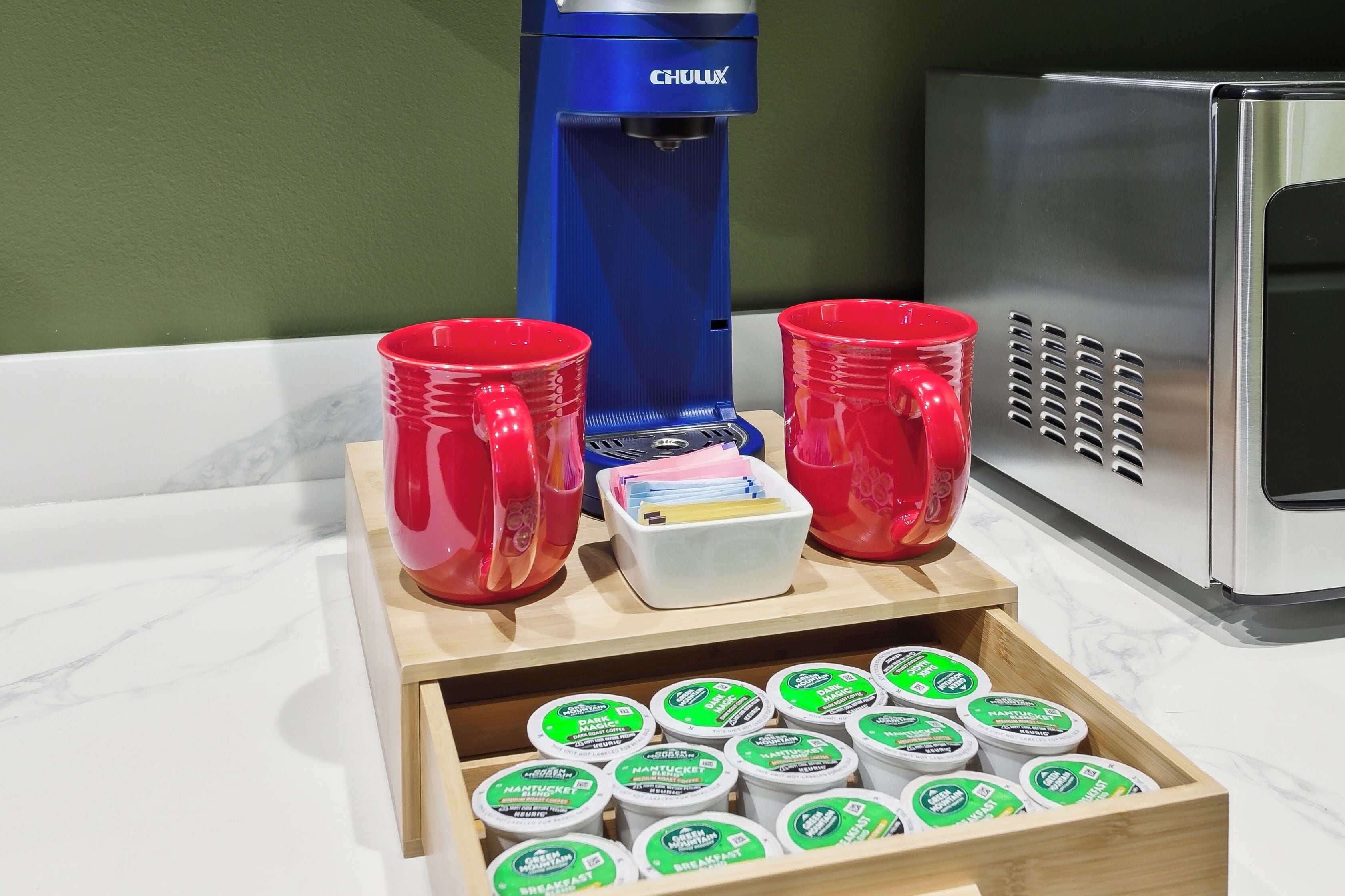 A kitchen counter with a coffee maker, red cups, and a drawer of yogurt containers.