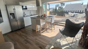 A modern kitchen with a central island, white cabinetry, and stainless steel appliances. There is a dining area with a table and chairs, and a balcony with outdoor seating. - Naturiste Cap d'Agde Villa Port Nature 187 (Agde)