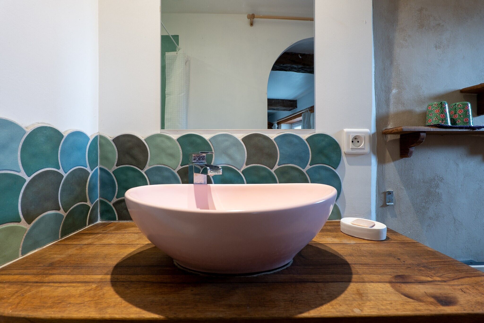 Un baño con un lavamanos rosa grande y redondo sobre una encimera de madera, un espejo encima y una pared de azulejos con un motivo verde y azul.