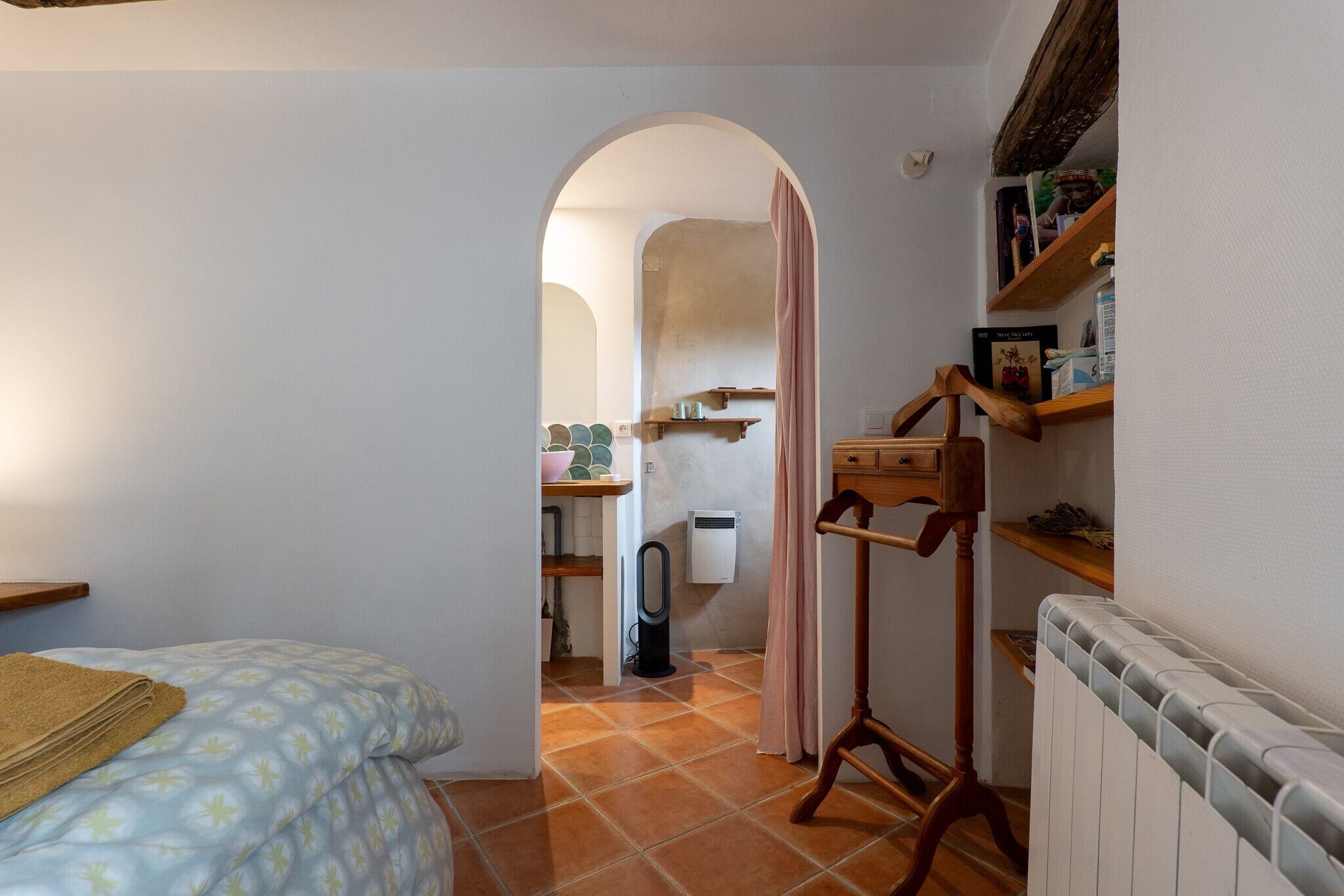 A compact room with a bed, a radiator, a wooden stand, and a shelf with books and decorative items.