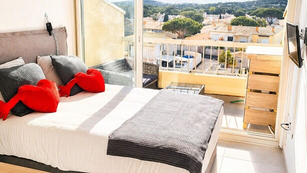 Une chambre à coucher avec un lit, une commode et une vue sur le paysage urbain, visible à travers une grande fenêtre.