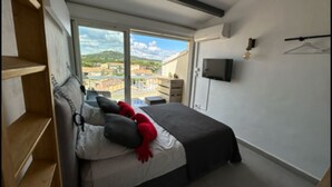 Une chambre à coucher avec un lit, un téléviseur et une vue sur le paysage urbain, visible à travers une grande fenêtre.