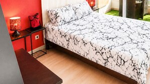 Een slaapkamer met een bed, nachtkastje, lamp, rode wand en raam met uitzicht op palmbomen.