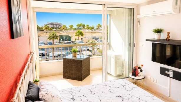 Een balkon met een rieten stoel, een klein tafeltje met een plant en een aan de muur bevestigde televisie.