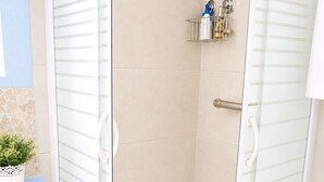 Douchecabine met glazen wanden en een witte deur, een klein plankje met toiletartikelen en een blauwe prullenbak.