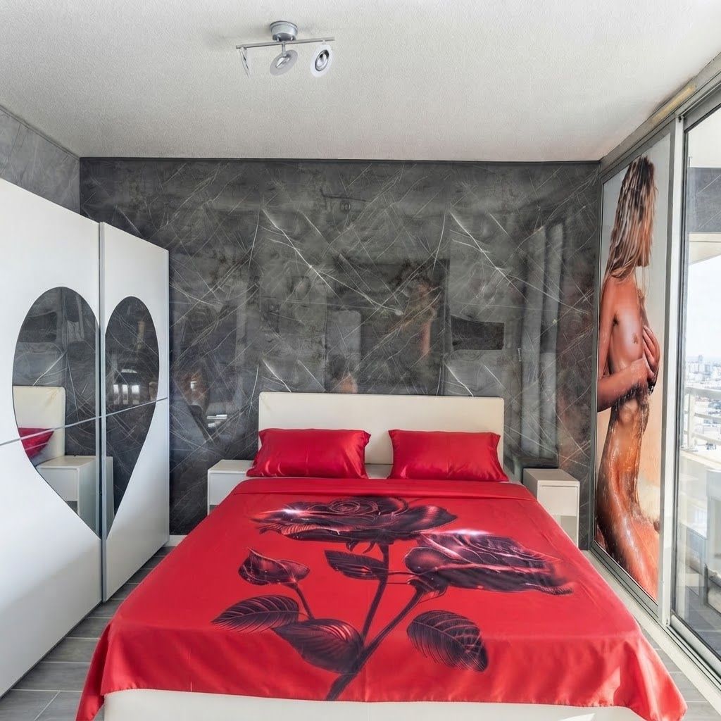 A modern bedroom with a large bed, a red floral bedspread, a city view through a large window, and a unique wall design.