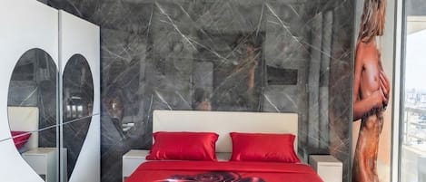 A modern bedroom with a large bed, a red floral bedspread, a city view through a large window, and a unique wall design.