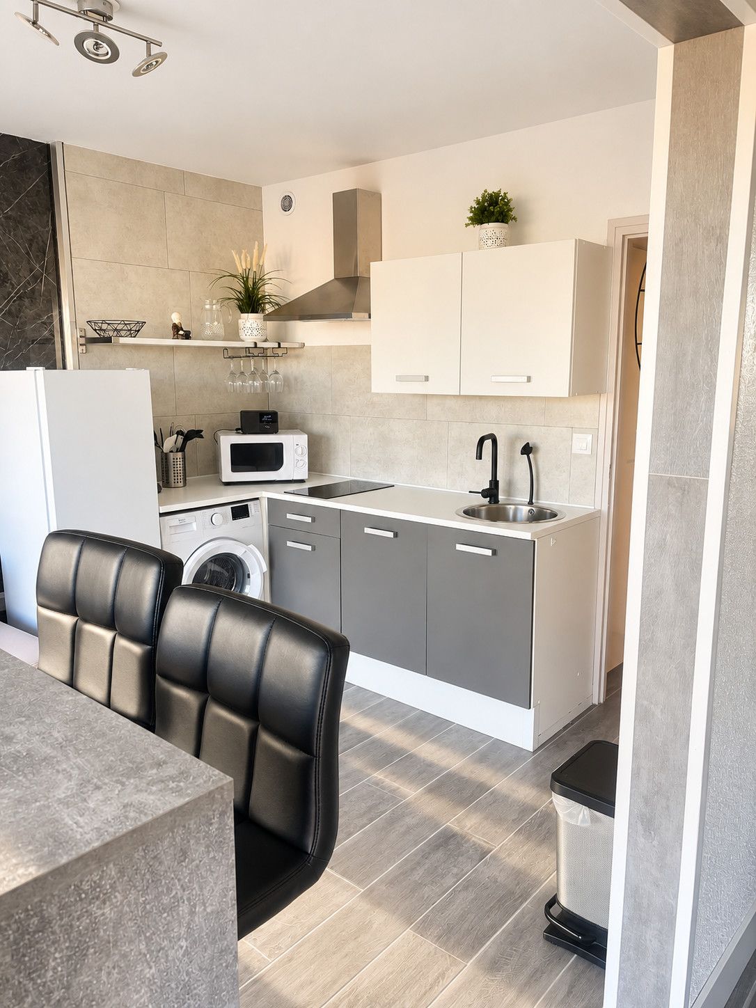 Una cocina moderna con lavadora integrada, microondas y lavamanos. Hay dos sillas negras en una mesa, y se ve un cubo de basura.