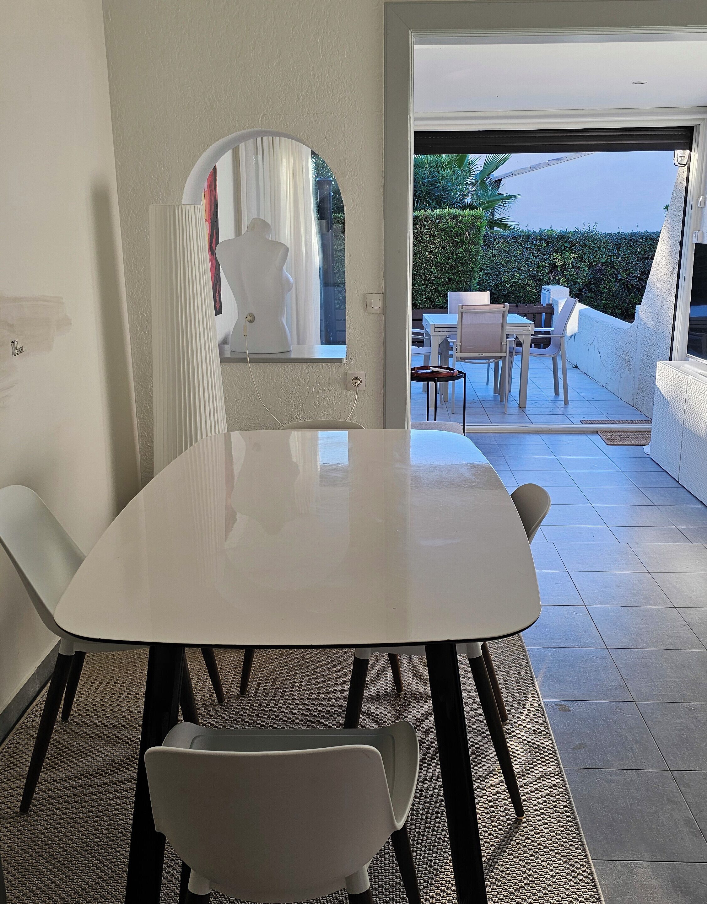 Un espace repas moderne avec une table et des chaises blanches, un miroir décoratif et une vue sur une terrasse extérieure équipée d’une table et de chaises.