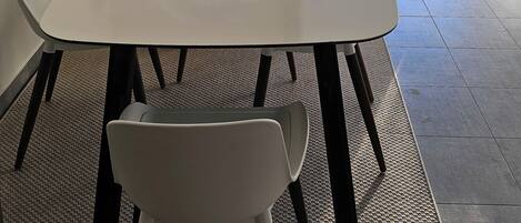 Un espace repas moderne avec une table et des chaises blanches, un miroir décoratif et une vue sur une terrasse extérieure équipée d’une table et de chaises.