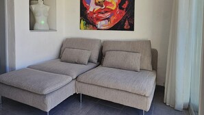 A modern living room with a grey sectional sofa, a colorful abstract painting, and a small arched window. - Naturiste Cap d'Agde Villa Héliovillage 20 (Agde)
