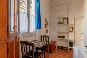Una habitación con una mesa de comedor de madera, sillas, un armario, una ventana con cortinas y una estantería blanca con marcos de fotos.