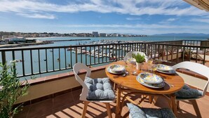 Ein Balkon mit Tisch für zwei Personen, Blick auf einen Yachthafen mit Booten und einer Stadtlandschaft im Hintergrund.