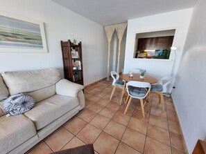 Ein Wohnzimmer mit einer Couch, einem Esstisch mit Stühlen, einem Bücherregal und einem Fenster mit Vorhängen.