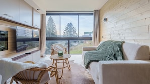 A modern living room with a large window, a flat-screen TV, a sofa, a chair, and a small table. - Lodge Belle Hutte - Proche des Pistes (La Bresse)