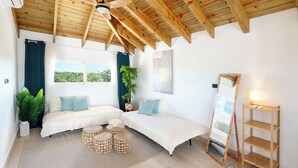 Una habitación con techo de madera, dos camas con ropa blanca, un ventanal con cortinas, una planta en maceta, un espejo y una estantería.