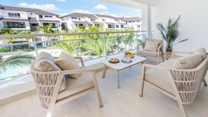 Un balcón con muebles de mimbre, una mesa con frutas y bebidas, y vista a una zona residencial con palmeras.