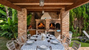 An outdoor patio with a brick fireplace, a round dining table set for six, and metal chairs. - VillaBerria, 9Host, Pool, BBQ, Jcuzzi, Oasis Palma (Marratxí)
