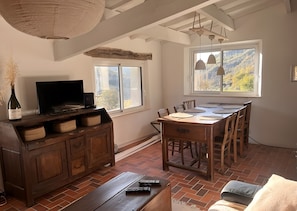 Un coin repas avec une table et des chaises en bois, un téléviseur posé sur un meuble en bois, et une fenêtre donnant sur des arbres.
