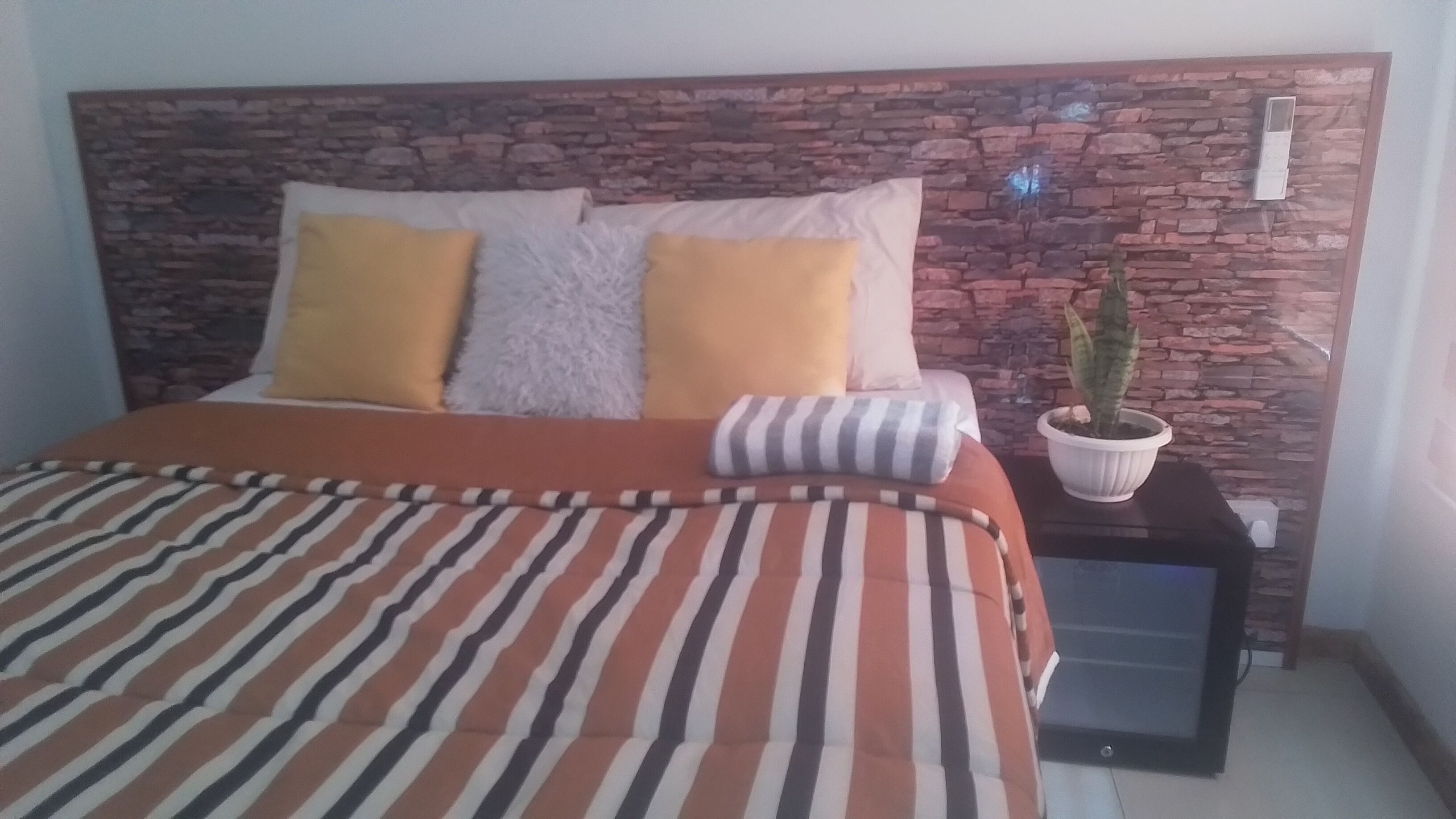 Un lit avec une literie à rayures, une tête de lit à motifs de pierre et une plante en pot posée sur une table de chevet.