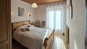 A bedroom with a wooden ceiling, a bed with a headboard, a bedside table, a chair, and a window with sheer curtains. - Comfortable Country House in Dordogne (Saint-Rabier)