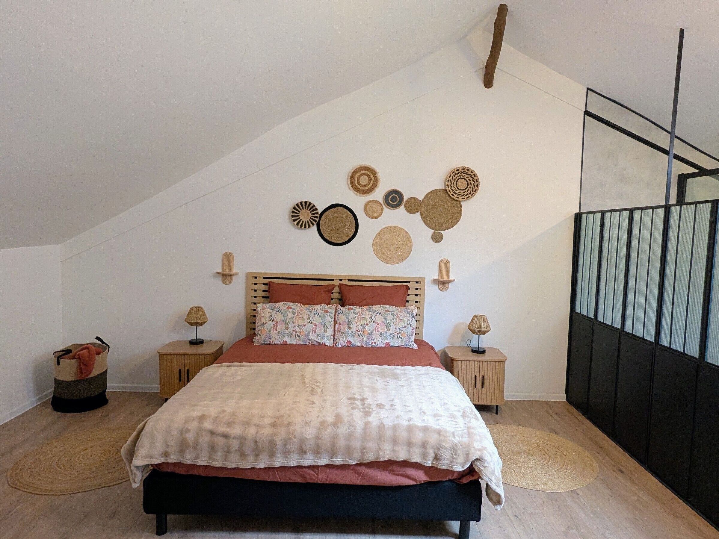 Un dormitorio con techo inclinado, una cama con colcha estampada, dos mesitas de noche con lámparas, y decoración en la pared que incluye cestas tejidas y un espejo.