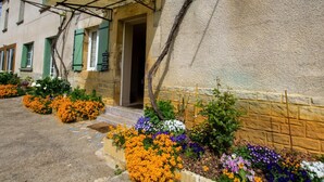 A building with a stone facade, green shutters, and a wooden pergola. There are flower beds with various colorful flowers in front of the entrance. - Cottage for 11 with parking, spa, pets allowed (Paulin)